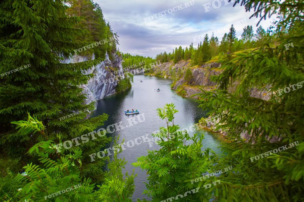 На фотографии изображена лодочная прогулка в мраморном карьере Рускеала в Карелии. Узкий водоём окружён высокими мраморными скалами и густыми хвойными лесами. Вода спокойная и прозрачная, что делает это место идеальным для отдыха на природе и наслаждения живописными видами. Рускеальский горный парк предлагает уникальные экскурсии и активные виды отдыха, привлекая туристов со всего мира.